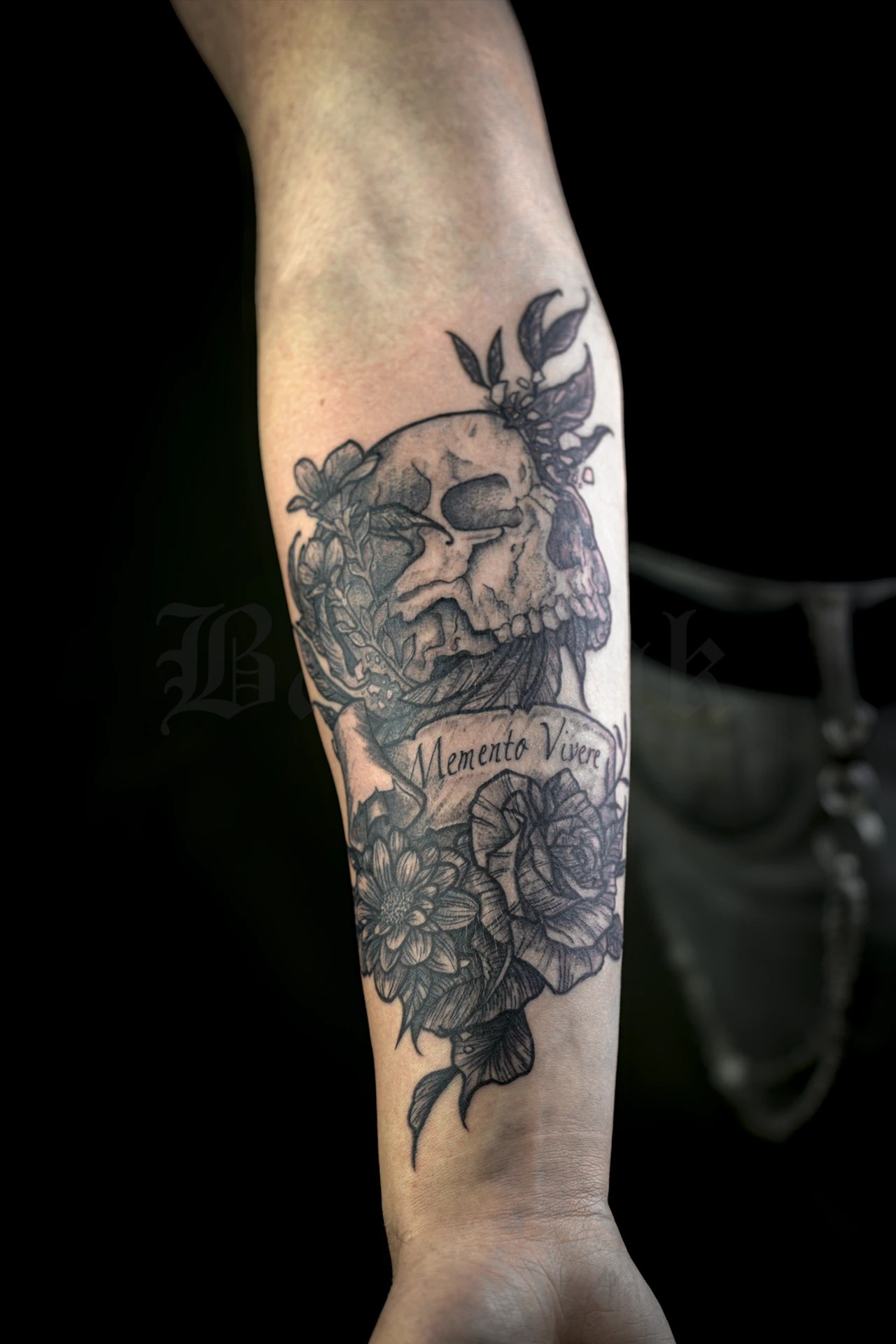 Tatouage noir et gris de crâne entouré de fleurs et de feuillages, avec inscription Memento Vivere