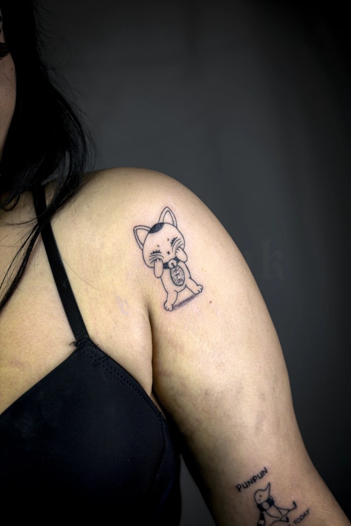 Petit tatouage de chat de style manga sur l’épaule, les pattes contre les joues, dessiné en traits noirs