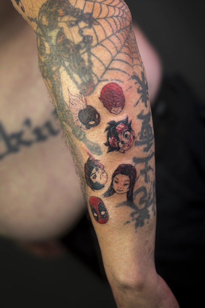 Petits portraits de personnages de comics en couleur sur l’avant-bras, dont Deadpool et Daredevil